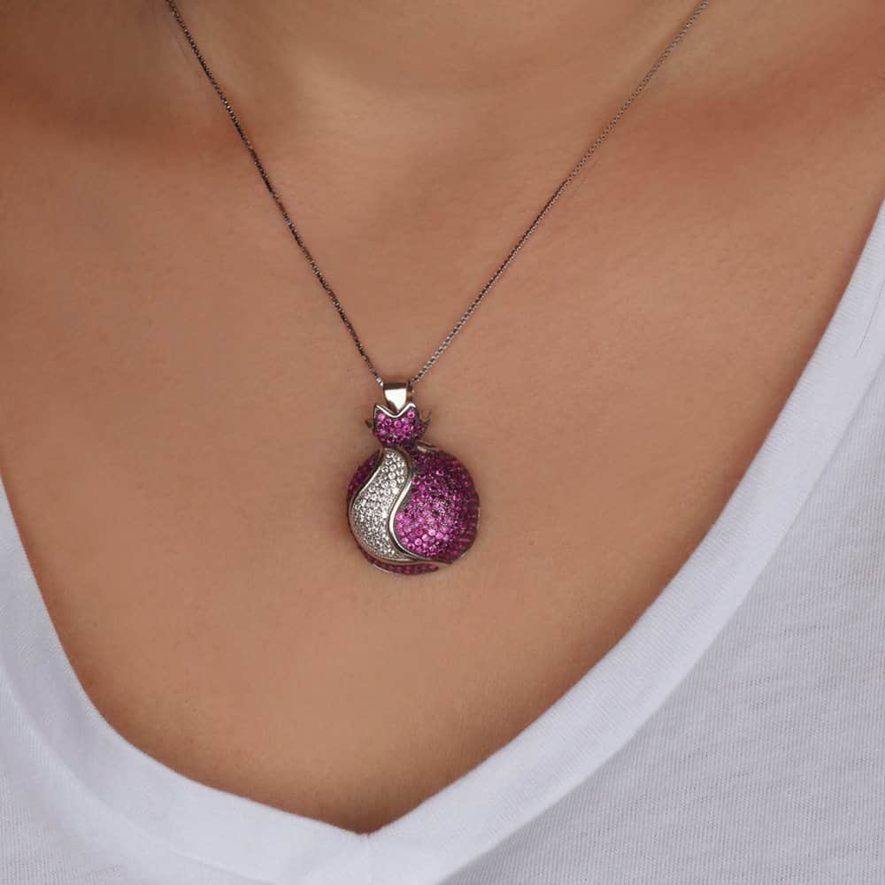A close-up of a person's neck and chest wearing a silver chain necklace with a pomegranate pendant. The pendant features a bright pink textured top section and a silver textured bottom section, set against the model's skin and white V-neck top.