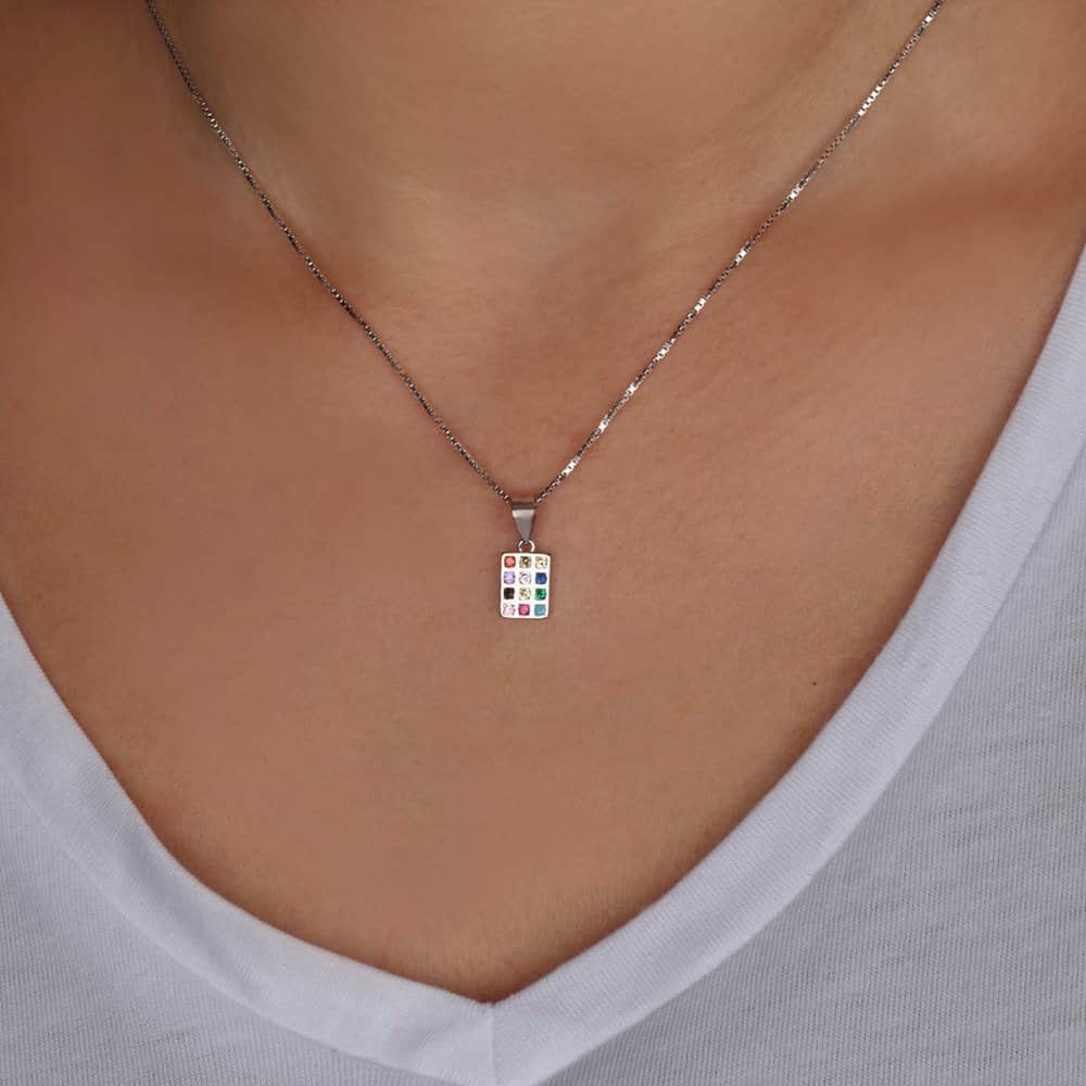 A silver-toned necklace with a small rectangular pendant featuring a colorful grid of tiny colored squares, worn on a model's neck over a white V-neck top.