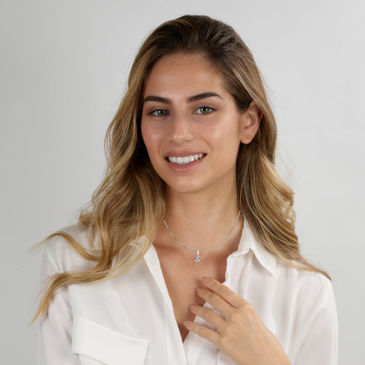 A smiling woman with blonde-highlighted hair wearing a delicate silver necklace with a small geometric pendant, paired with a white collared blouse, set against a plain light gray background.