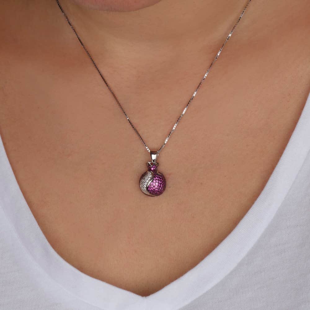 A close-up of a person wearing a silver chain necklace with a small round pomegranate pendant featuring a textured pink and silver design.