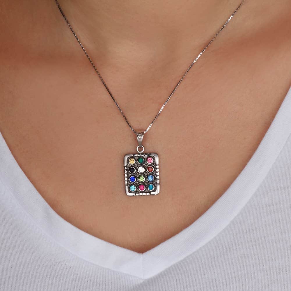 A silver-toned rectangular Hoshen Twelve Tribes necklace with a colorful, mosaic-like pattern of small gemstones, worn on a model's neck over a white V-neck top.